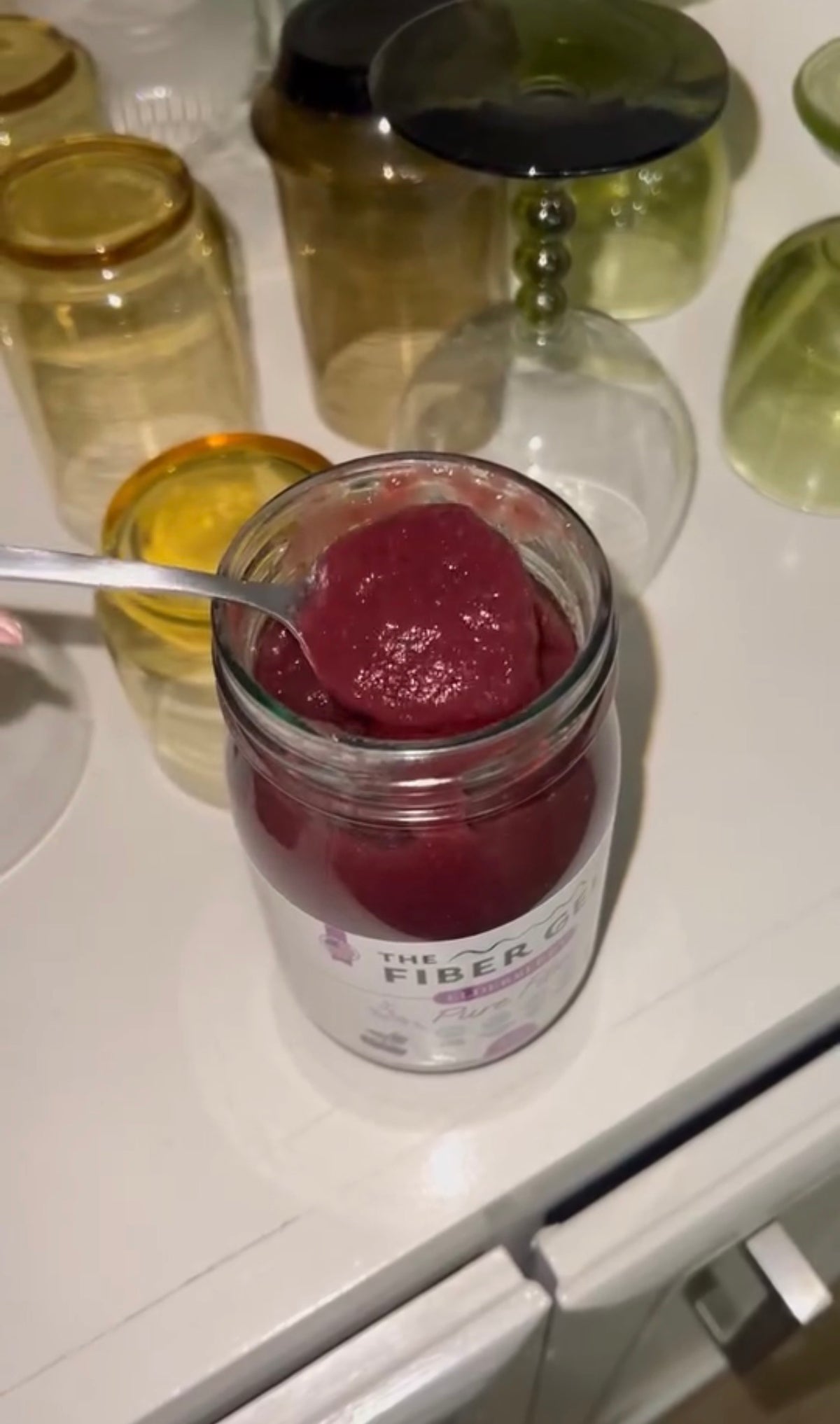 Jar of red substance with a spoon on a white surface with glass containers in the background